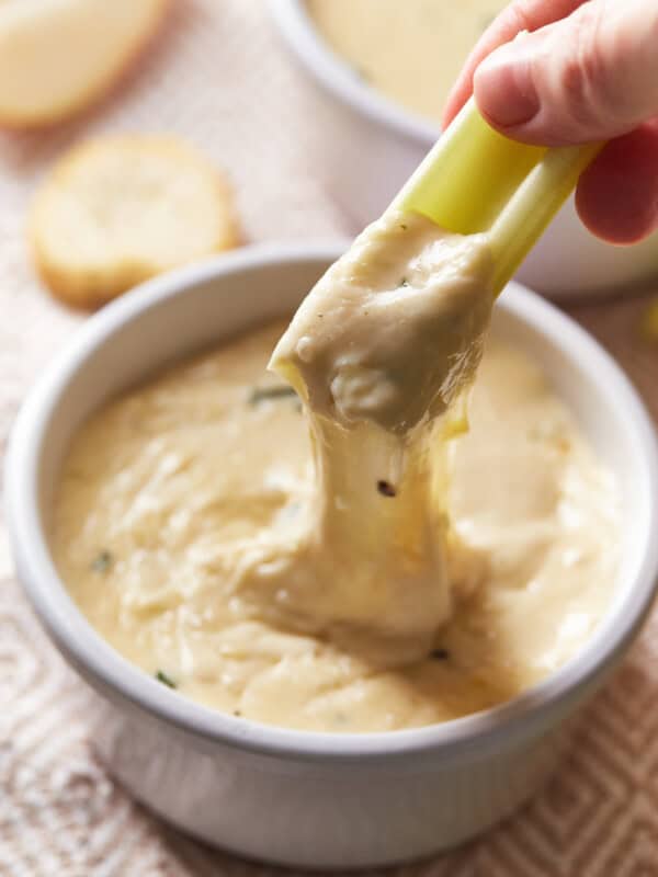 a hand pulling a piece of celery out of white wine fondue in a white bowl, creating a cheese pull.