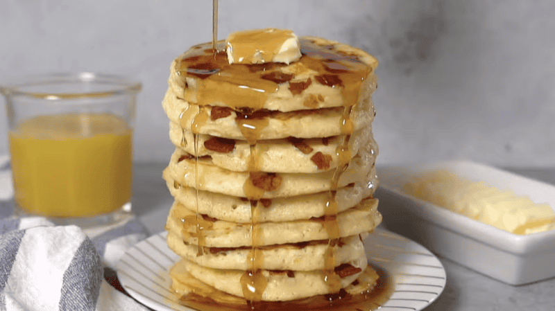 pouring maple syrup over a stack of bacon pancakes topped with butter.