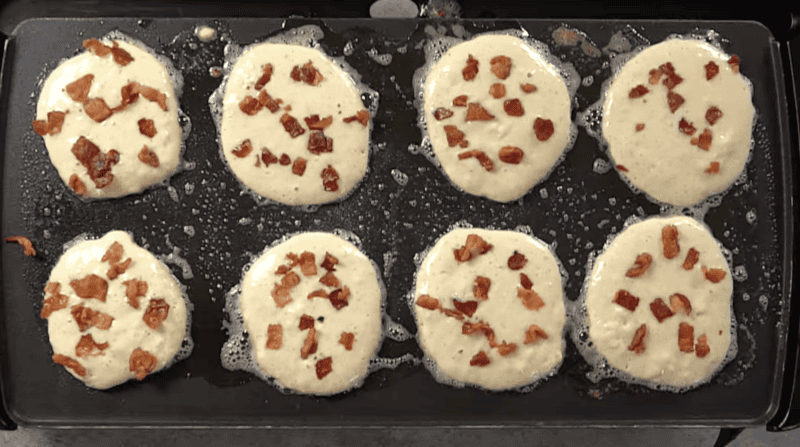 8 pancakes topped with bacon bits on a griddle.