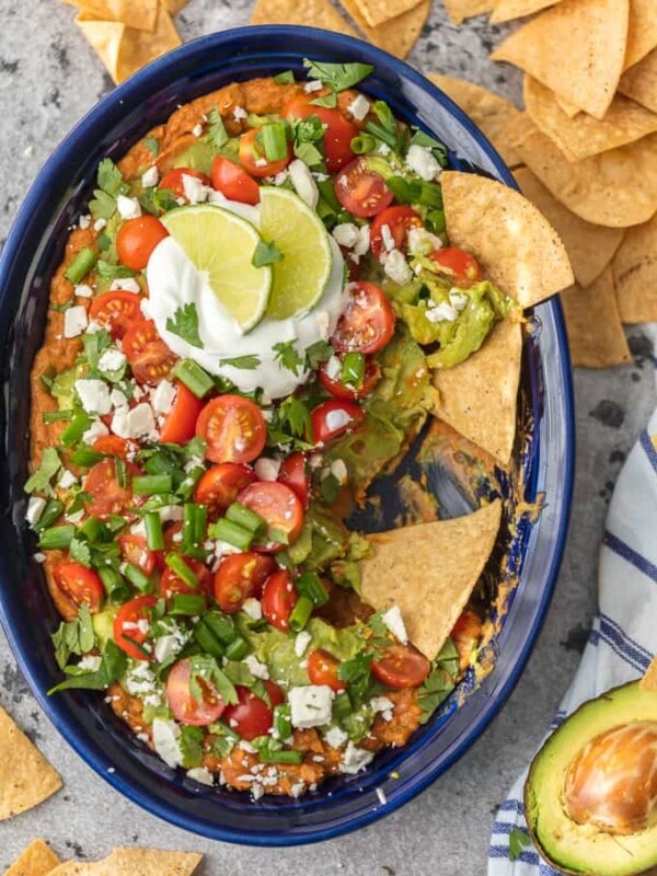 This Layered Bean Dip Recipe is one of our favorite ways to tailgate! Guacamole Bean Dip has layers of guacamole, spicy refried beans, tomatoes, onions, and cheese. This Mexican Bean Dip is SO delicious and made in minutes! It doesn't get better than Layered Bean Dip for any and every celebration! Learn how to make bean dip with this recipe.