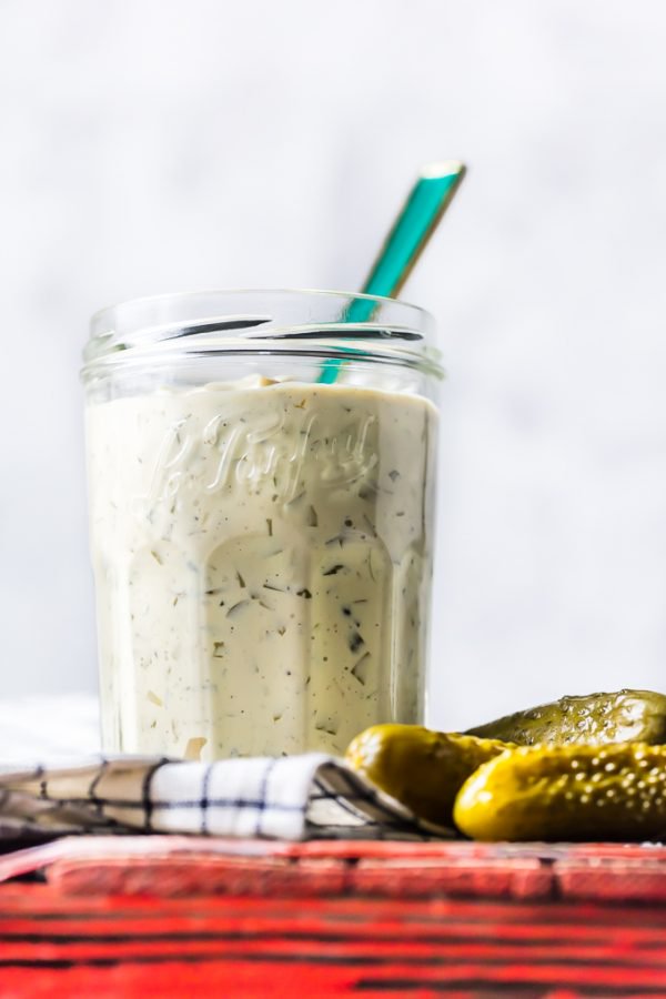 A jar of homemade tartar sauce on a table next to a pickle.