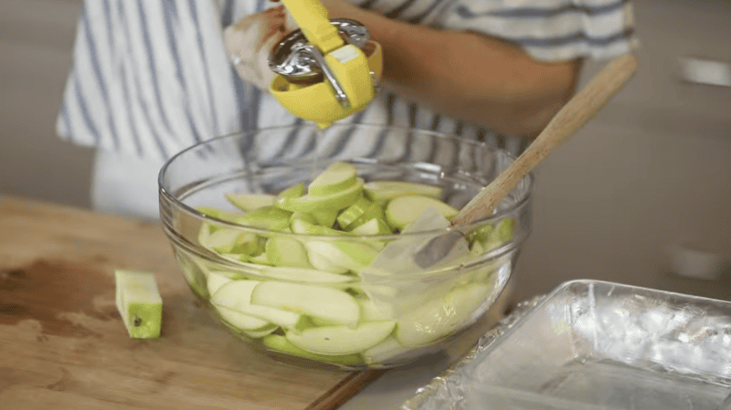 squeezing lemon juice over sliced apples in a glass bowl with a rubber spatula.