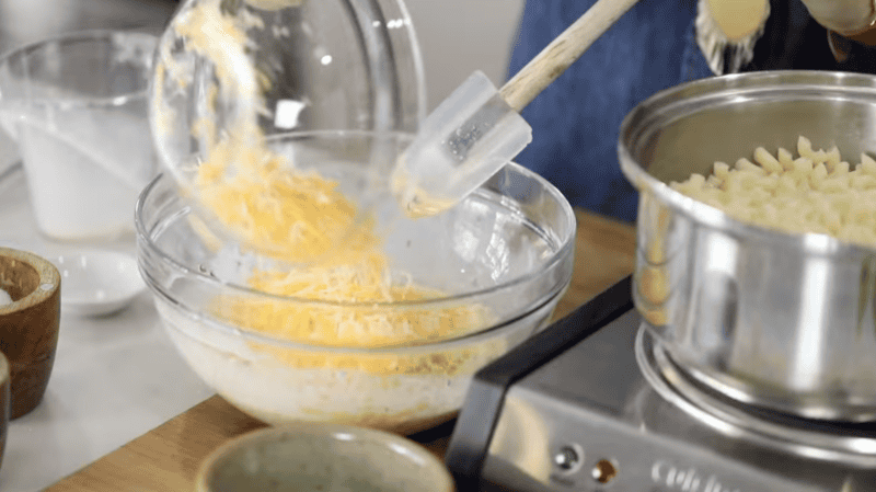 adding shredded cheese to mac and cheese sauce mixture in a glass bowl.