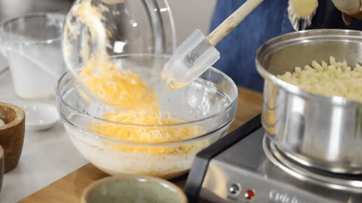 adding shredded cheese to mac and cheese sauce mixture in a glass bowl.