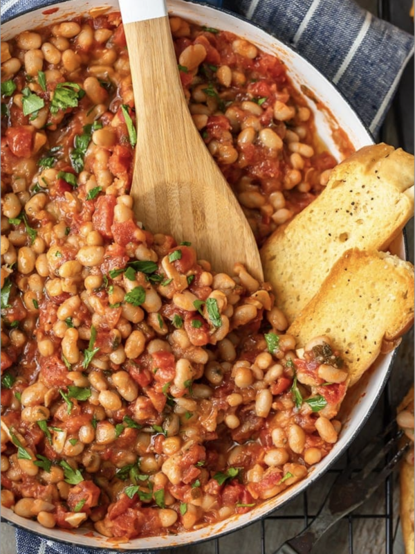 A pan full of white beans in tomato sauce, being stirred with a wooden spoon.