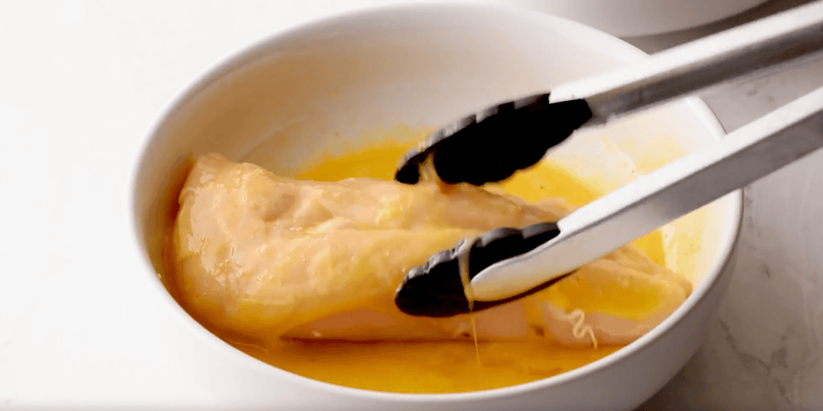 using tongs to dip a chicken breast in egg.