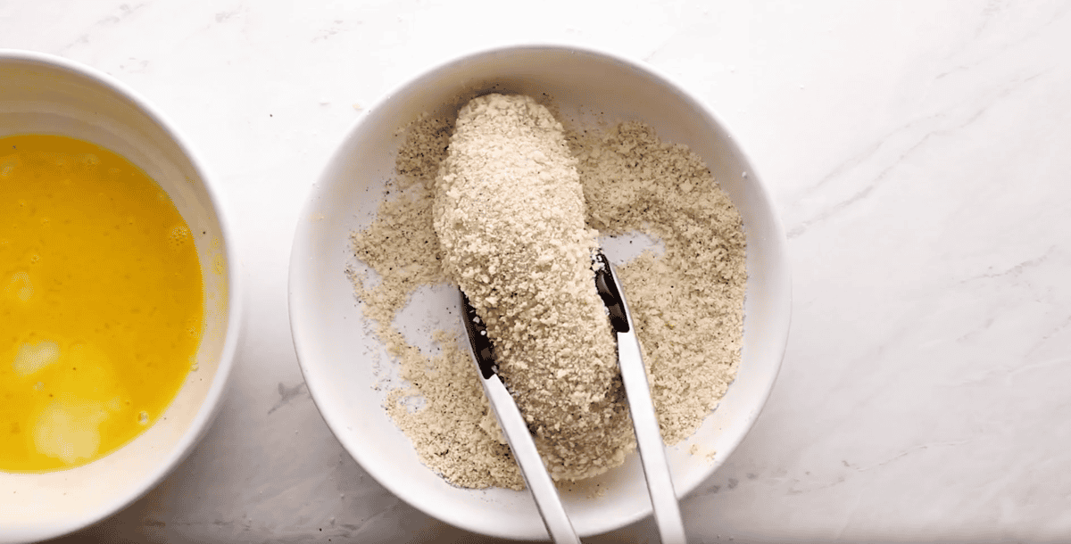 lifting a panko-coated chicken breast from a shallow white dish with tongs.