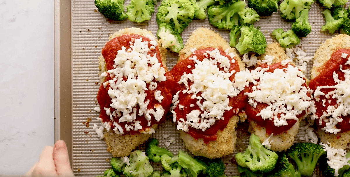 shredded mozzarella cheese and marinara sauce over baked breaded chicken breasts and broccoli on a baking sheet.