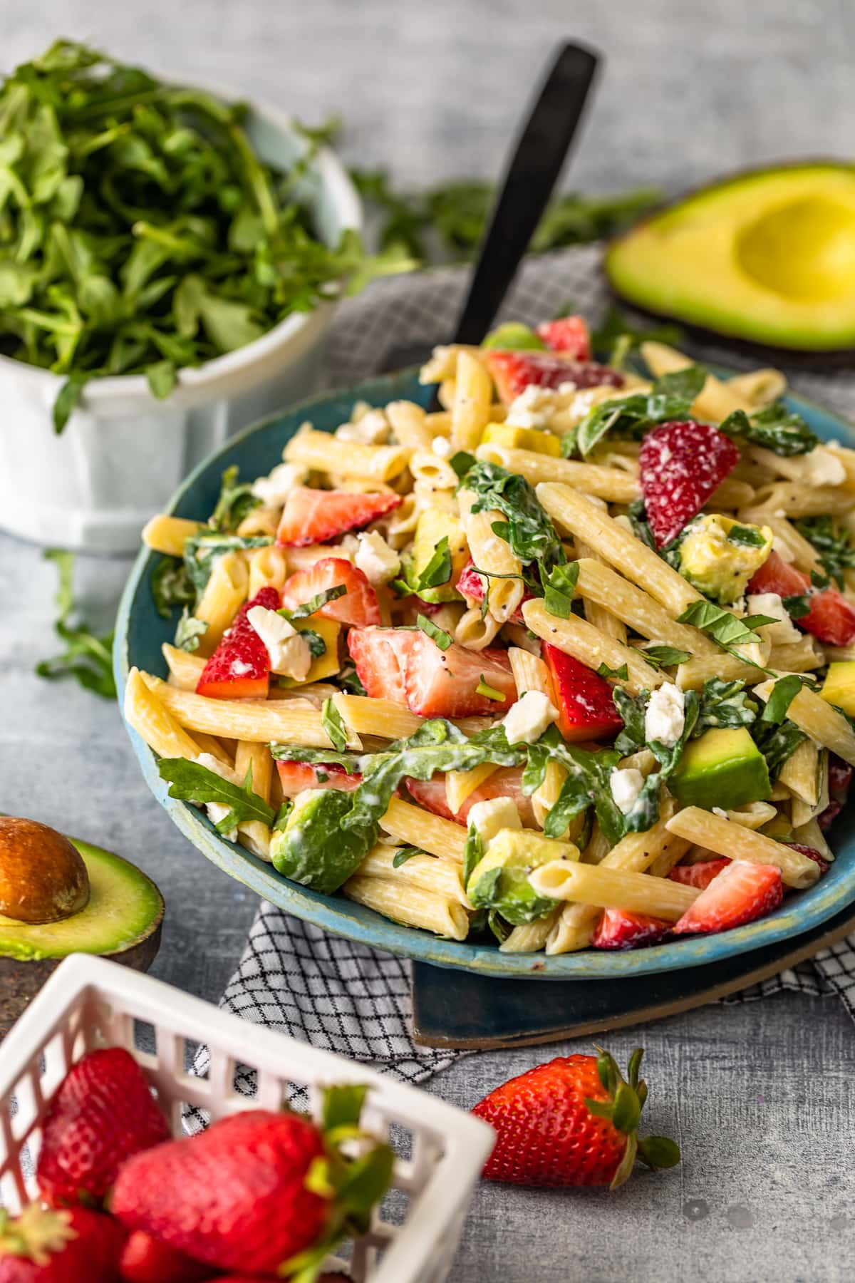 bowl of pasta salad surrounded by avocados, strawberries, and arugula