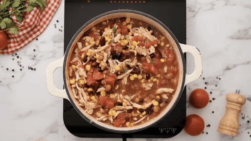 a dutch oven filled with tomatoes, beans, corn, and chicken.