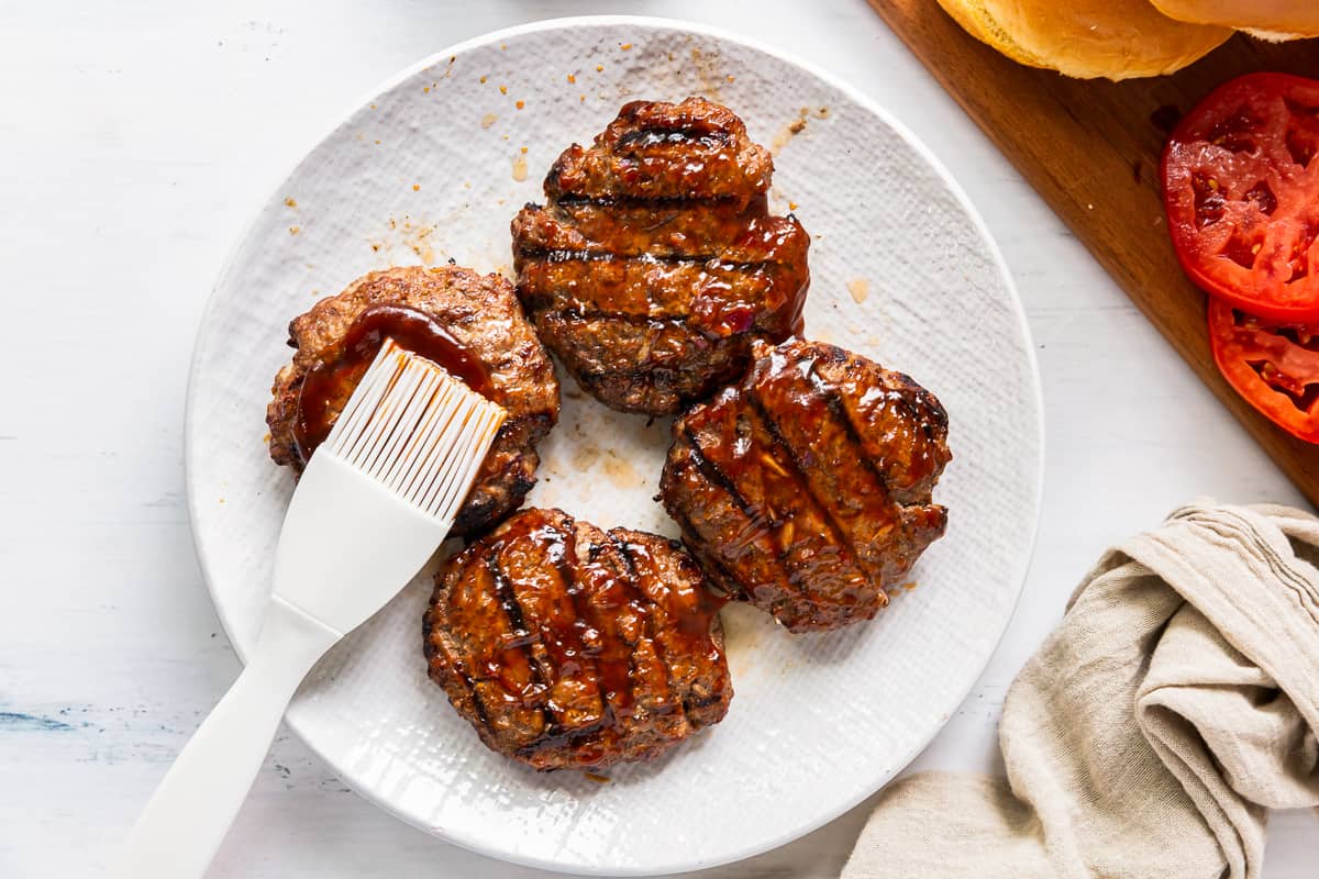 brushing grilled burger patties with bbq sauce on a plate.