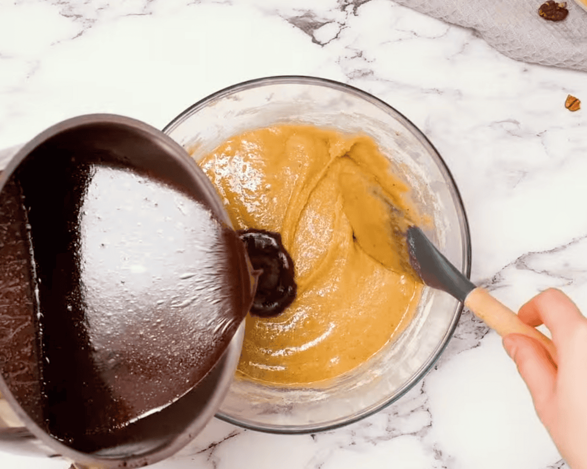 A saucepan pouring a hot chocolate mixture into the batter while a spatula is ready to mix it in.