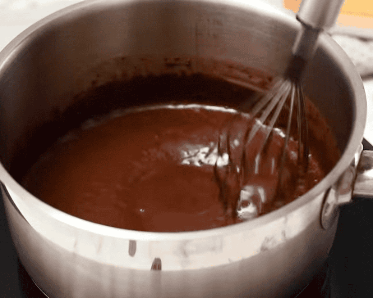 A saucepan on the stove with glossy chocolate frosting being whisked as it thickens.