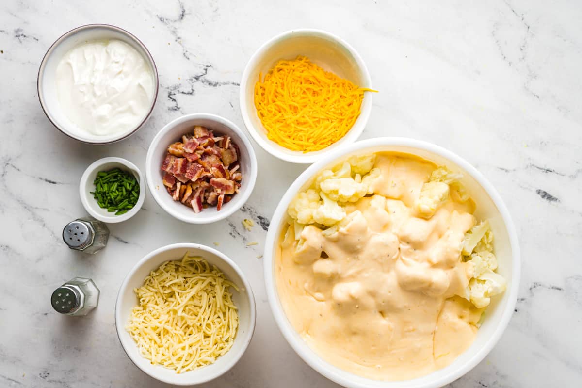 A bowl of cauliflower covered in a cheese sauce, surrounded by bowls of other ingredients.