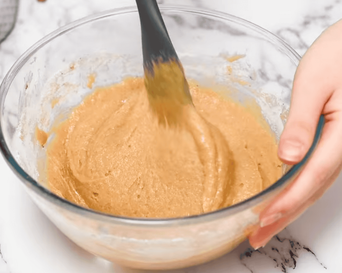 A hand stirring a thick, golden batter in a glass bowl with a silicone spatula.