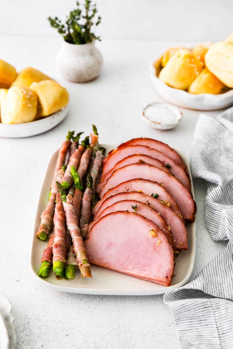 slices of brown sugar ham and prosciutto wrapped asparagus spears on a white serving tray