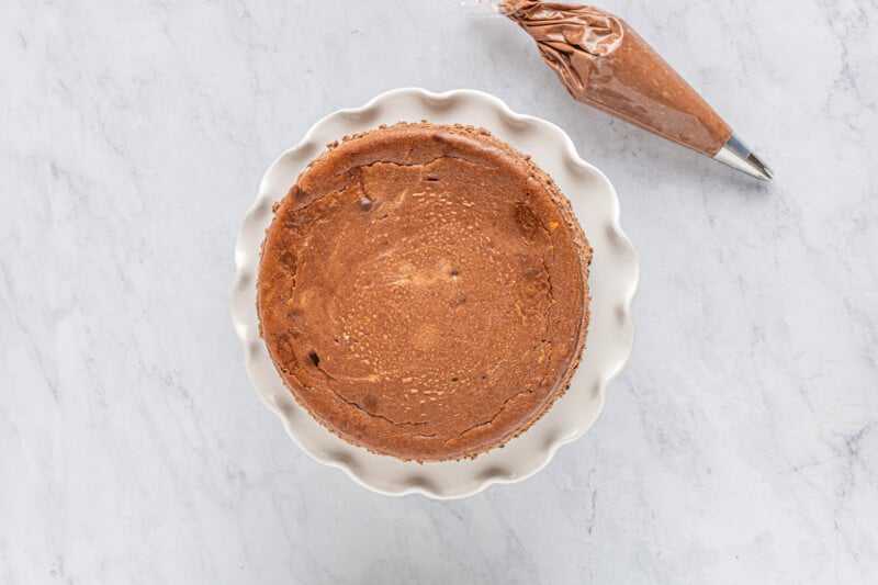 chocolate cheesecake on a white cake stand with chocolate frosting in a piping bag