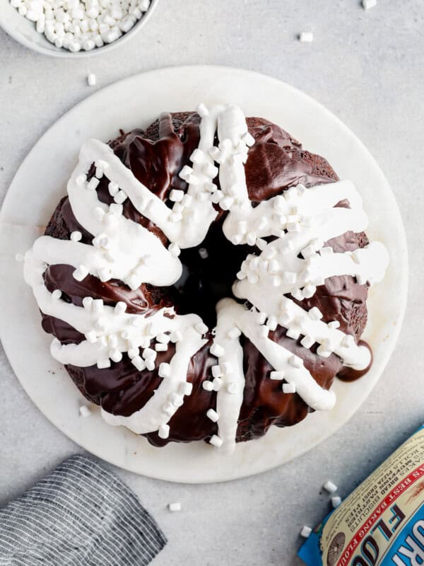 overhead image of chocolate bundt cake topped with chocolate ganache, marshmallow creme, and mini marshmallows on a white serving tray before slicing