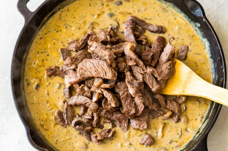 adding cooked beef strips to sauce in a skillet with a wood spoon