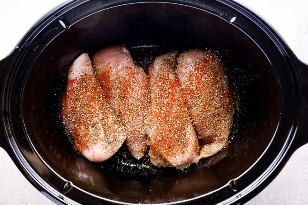 4 raw chicken breasts in the bottom of a Crockpot, covered in a mix of spices.