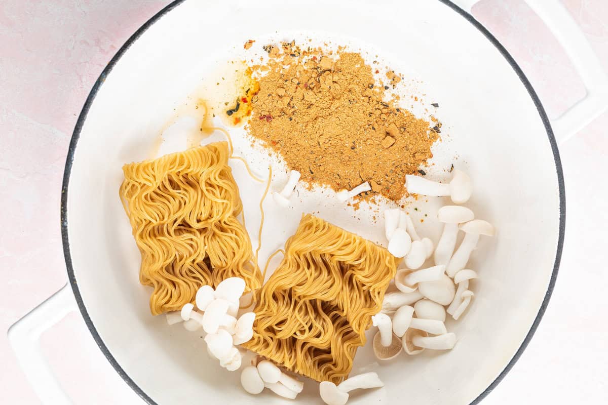 2 ramen noodle bricks in a white soup pot with seasoning and mushrooms.