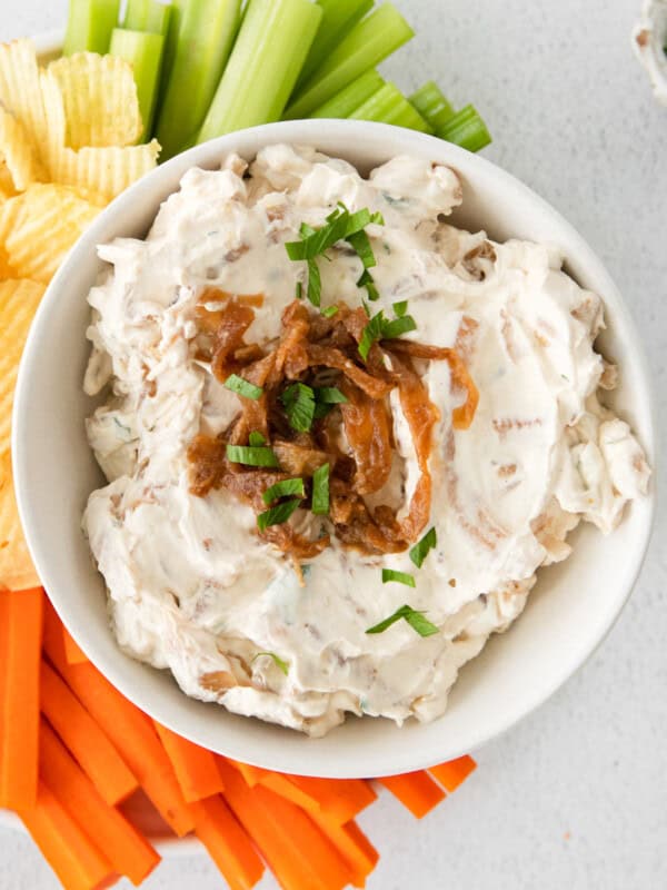 overhead image of french onion dip in a white bowl on a serving platter surrounded by chips, celery sticks, and carrot sticks