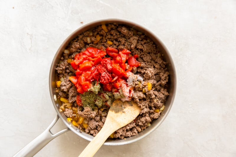 seasonings and tomatoes added to ground beef mixture in a skillet with a wood spoon