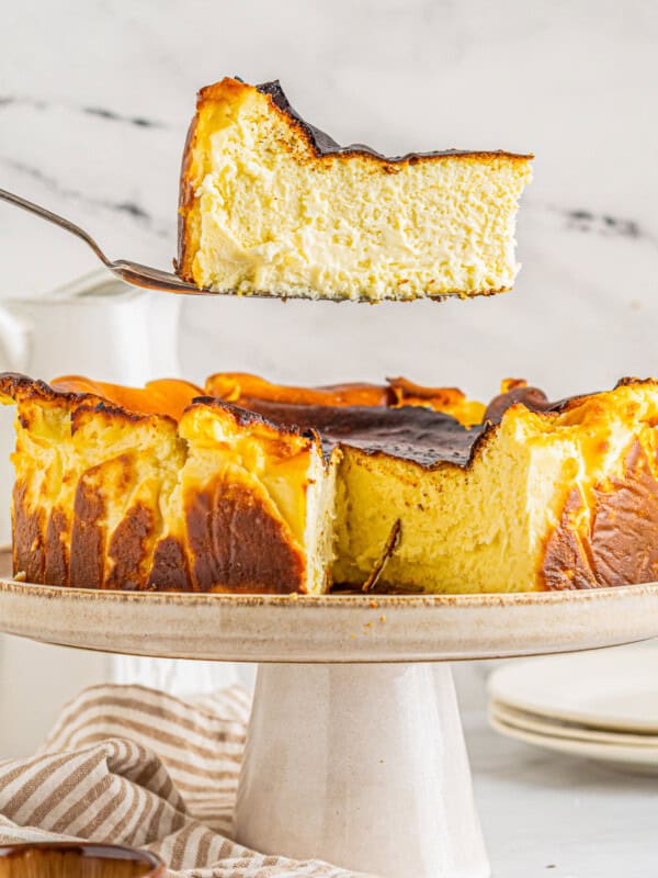 a slice of basque cheesecake on a cake server hovering above a cheesecake on a white cake stand.