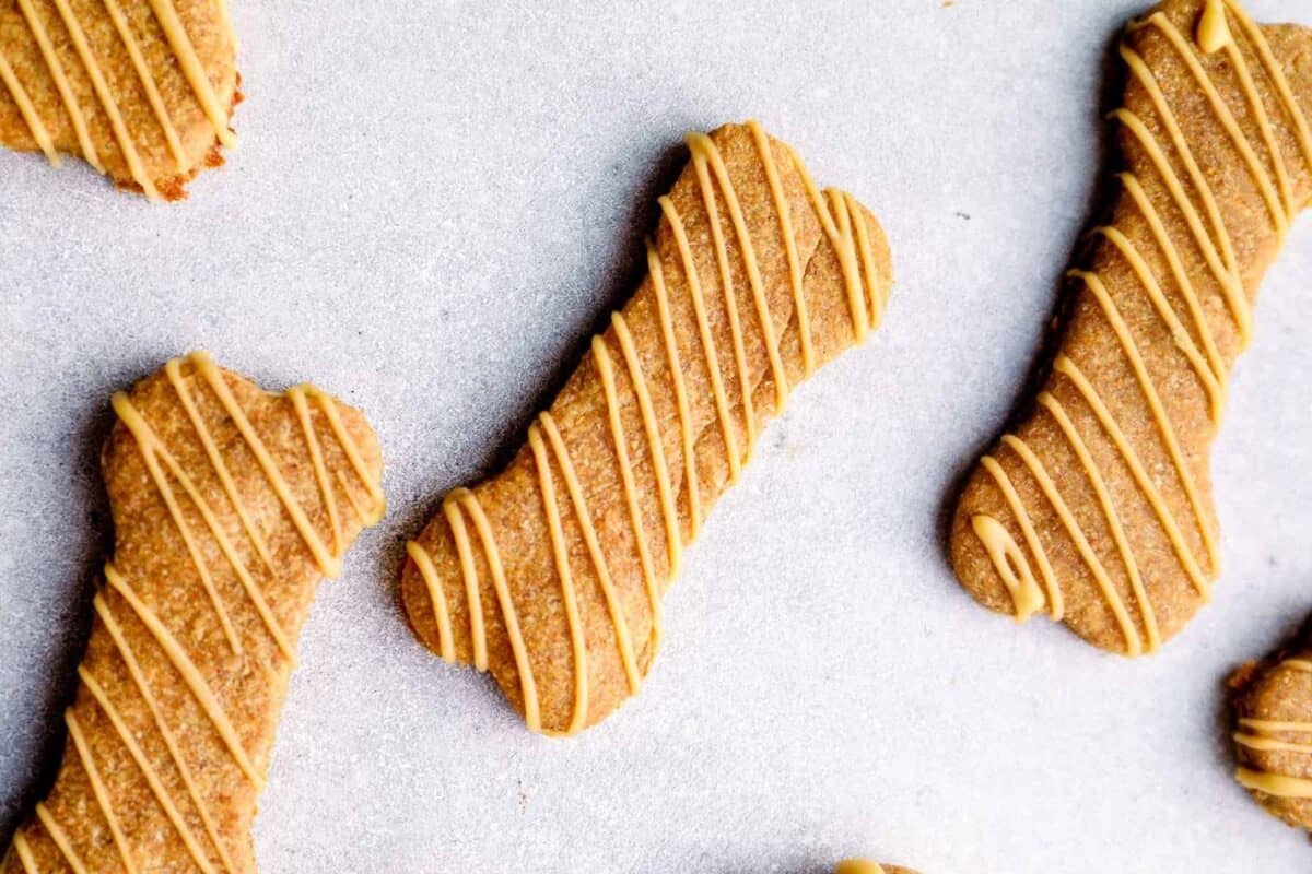 A close-up of homemade bone-shaped dog treats with a golden-brown color, drizzled with peanut butter frosting, placed on a parchment-lined surface.