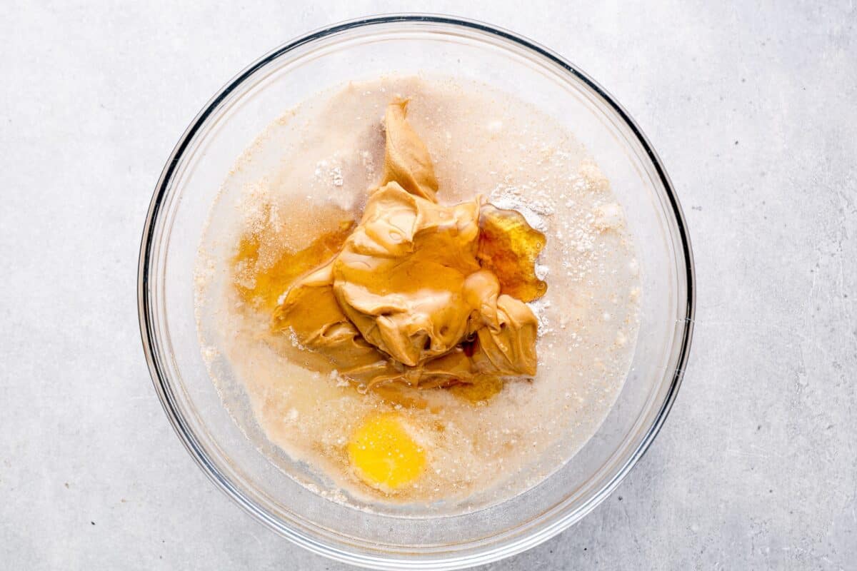 A glass mixing bowl with ingredients for dog treats, including peanut butter, honey, flour, egg, and water, before being mixed into a dough.