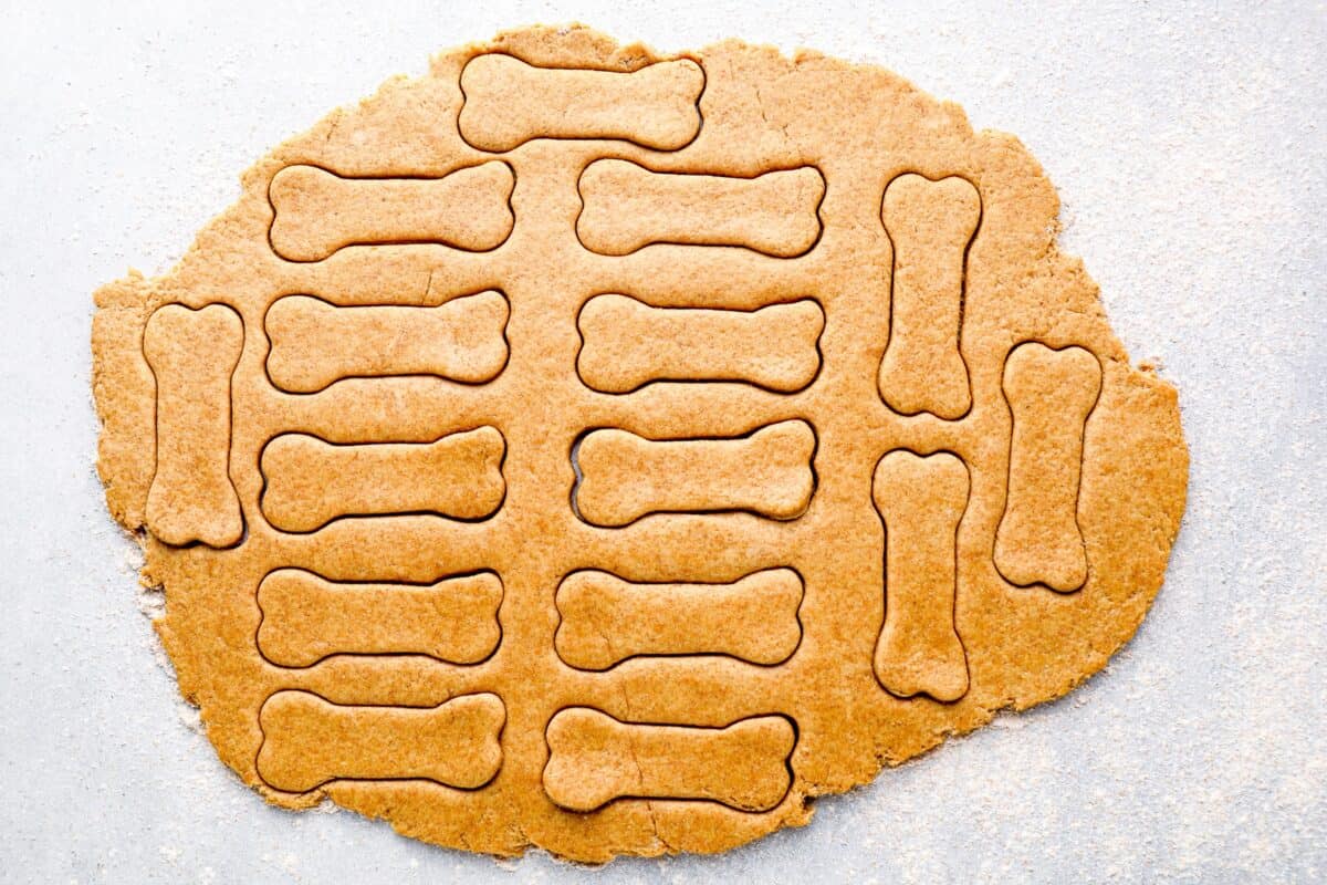 A large rolled-out sheet of dough with multiple bone-shaped dog treats cut out, still attached to the unbaked dough.