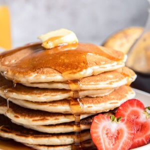 close up of a tall stack of buttermilk pancakes on a white plate with butter, syrup, and a halved strawberry.