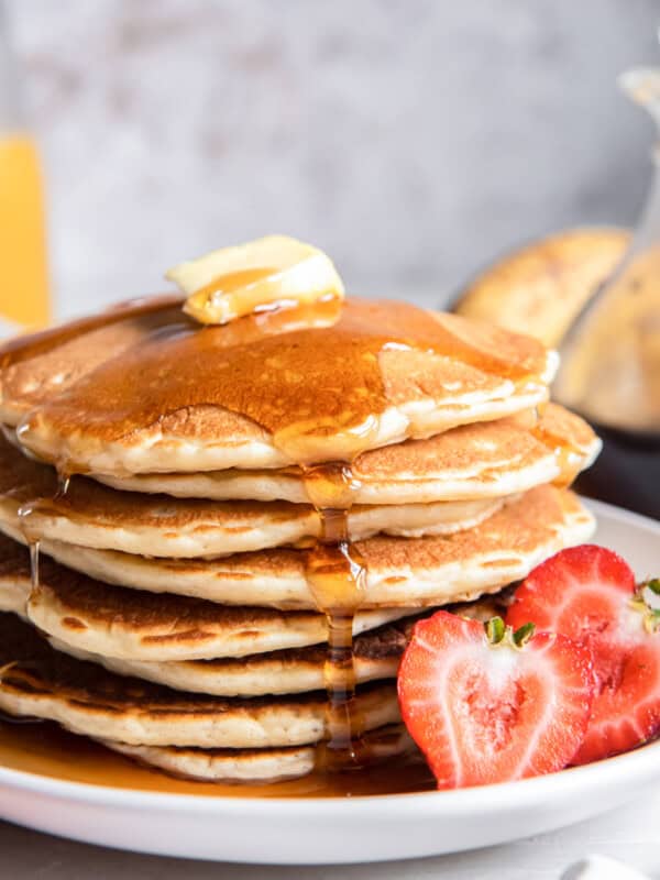 close up of a tall stack of buttermilk pancakes on a white plate with butter, syrup, and a halved strawberry.