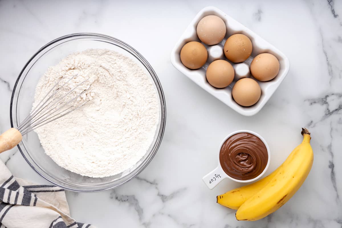 a whisk in a bowl of dry ingredients for nutella banana bread next to a half-dozen eggs, bananas, and nutella.