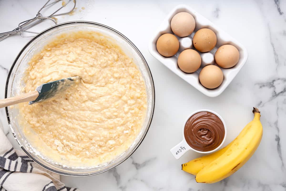 a spatula stirring nutella banana bread batter in a glass bowl next to a half-dozen eggs, bananas, and nutella.