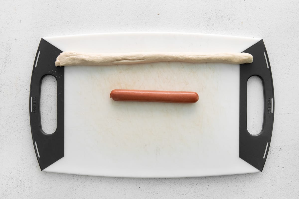 a hot dog and a piece of dough lined up on a cutting board