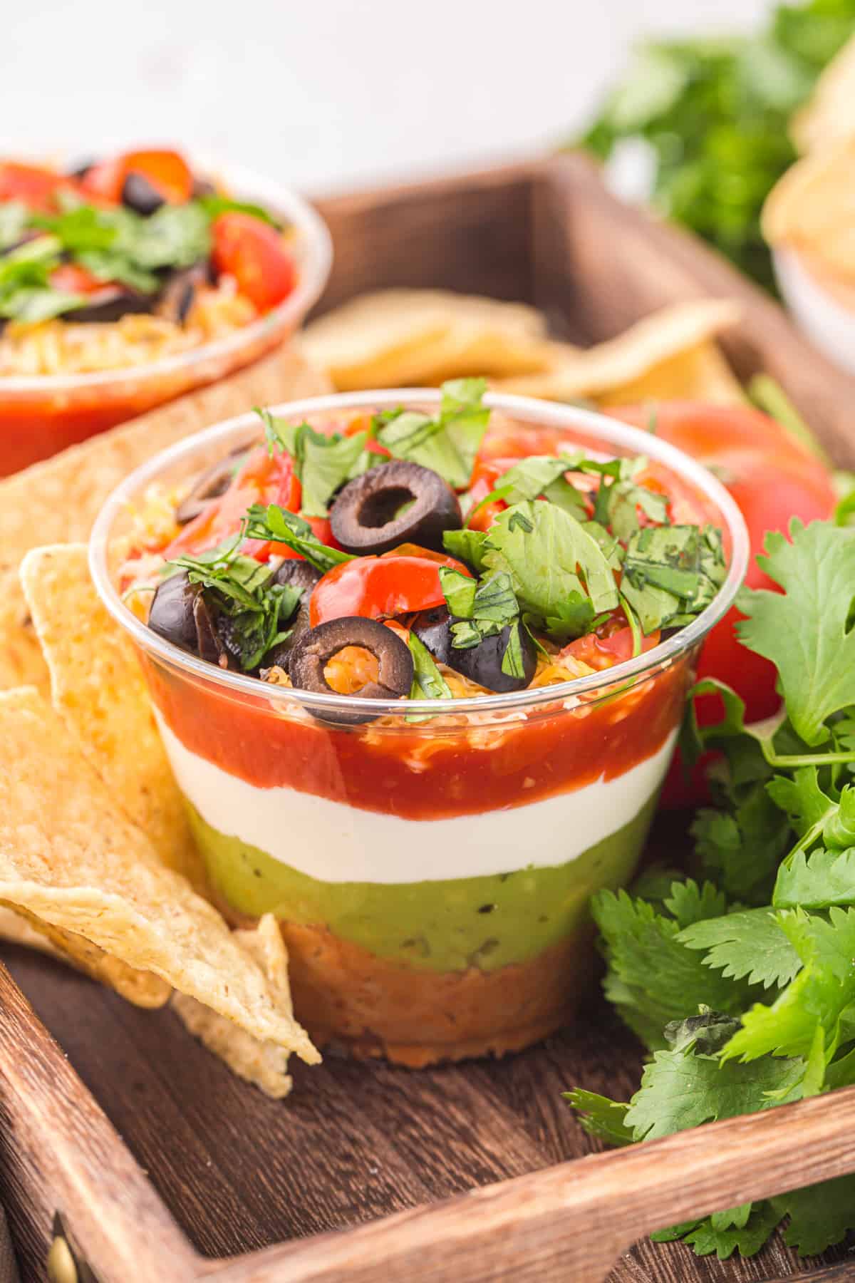 a 7 layer dip cup on a wooden tray with tortilla chips.