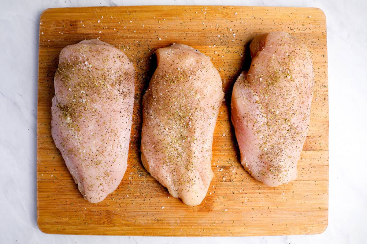 3 seasoned raw chicken breasts on a wooden cutting board.