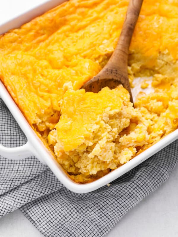 a wooden spoon scooping corn casserole from a white baking dish.