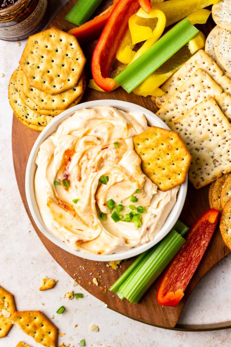 a board of crackers and veggies, with a bowl of pepper jelly cream cheese dip