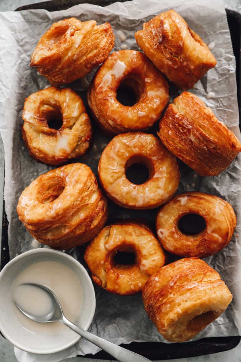 10 cronuts on a baking sheet with glaze in a white bowl with a spoon.