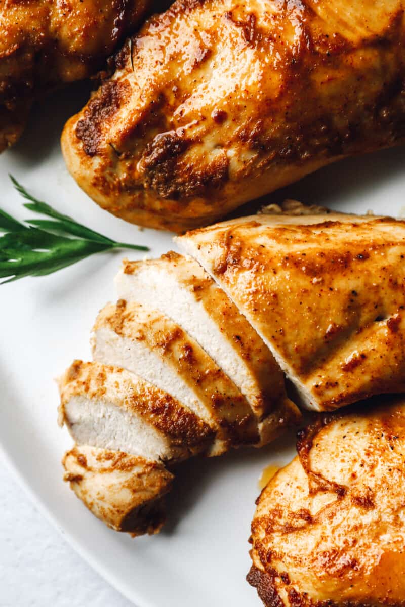 1 partially sliced chicken breast on a white platter next to 3 baked marinated chicken breasts.