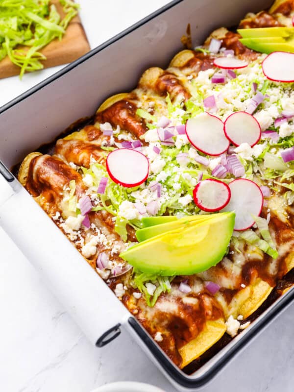 chicken enchiladas in a rectangular baking pan topped with lettuce, radish, onion, and avocado.