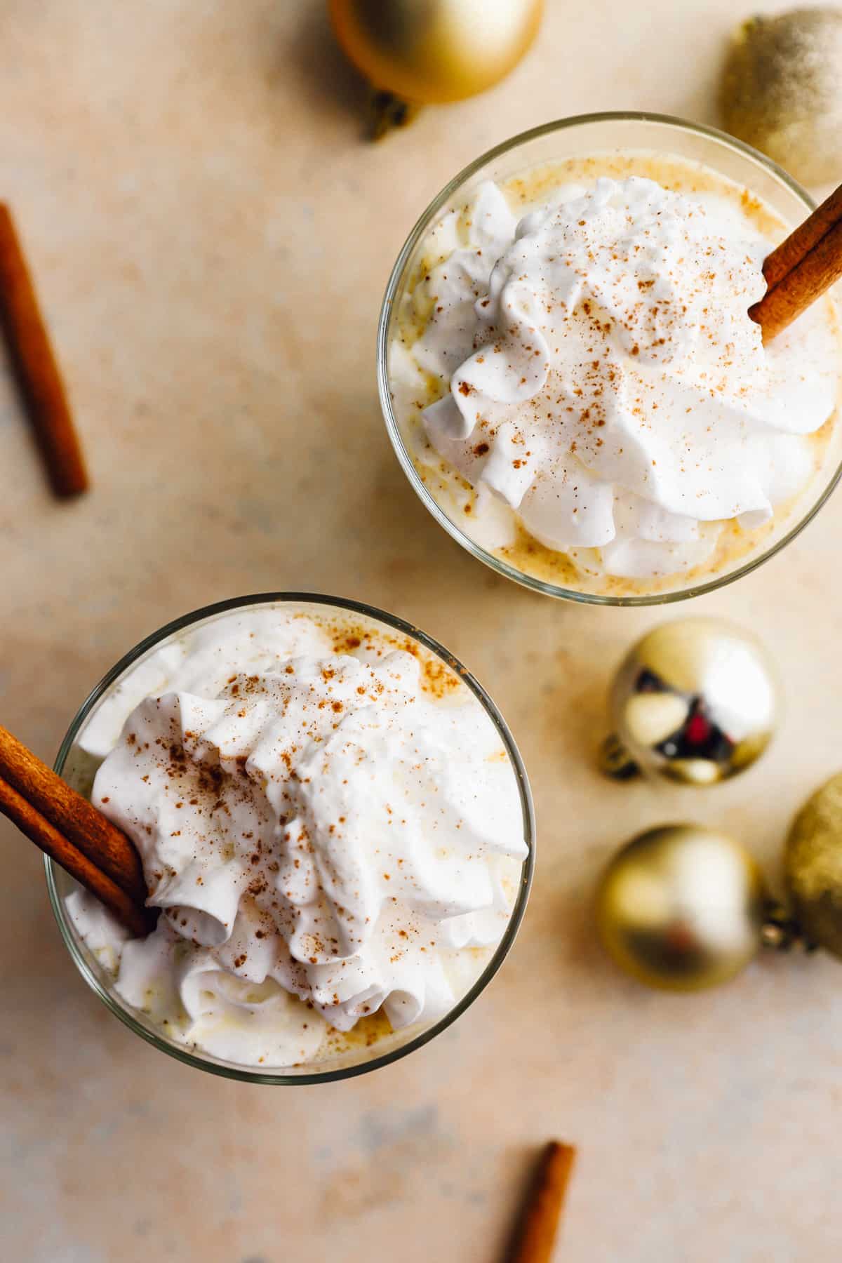 overhead view of 2 glasses of  eggnog with whipped cream and cinnamon sticks.