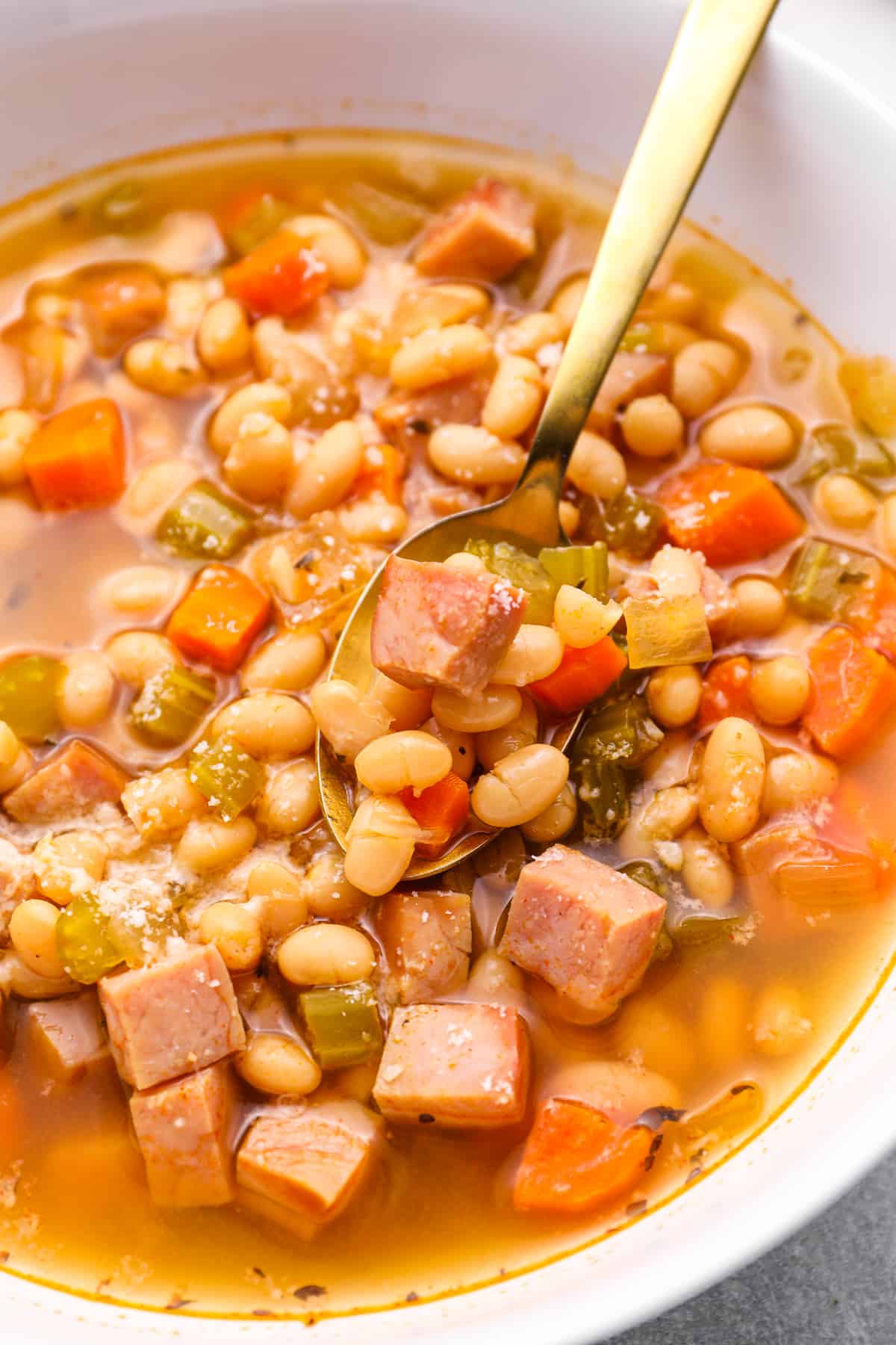 close up of ham and bean soup in a white bowl with a spoon.