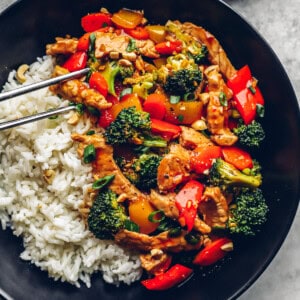 metal chopsticks grabbing a piece of pork from pork stir fry with white rice in a black bowl.