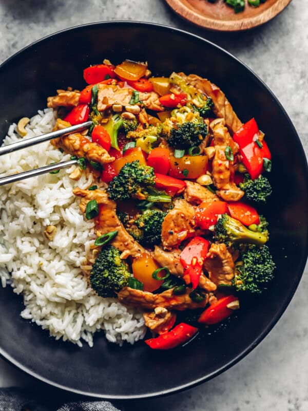 metal chopsticks grabbing a piece of pork from pork stir fry with white rice in a black bowl.