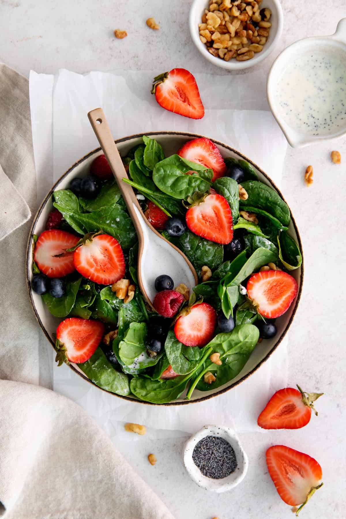 overhead view of spinach berry salad filled with strawberries, blueberries, raspberries, walnuts, and goat cheese, in a beige bowl with a serving spoon.