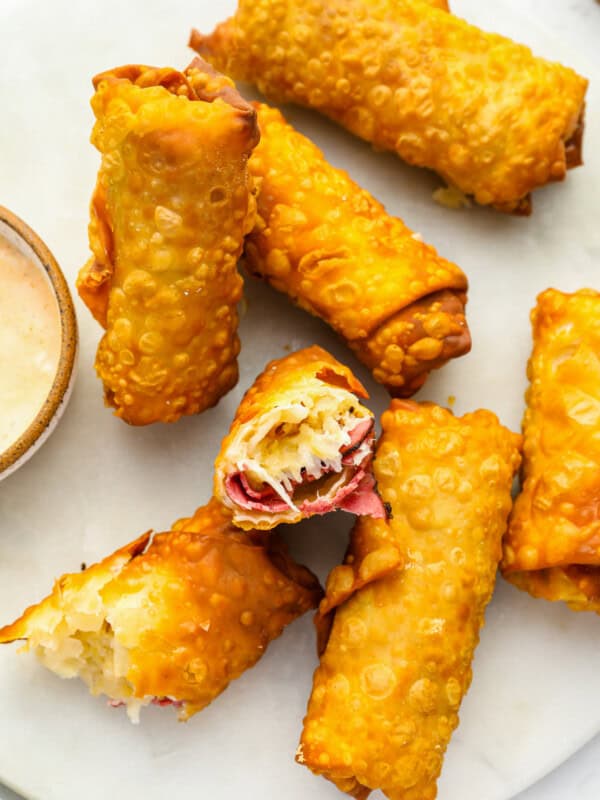 6 reuben egg rolls on a white plate, one broken in half to expose the filling.