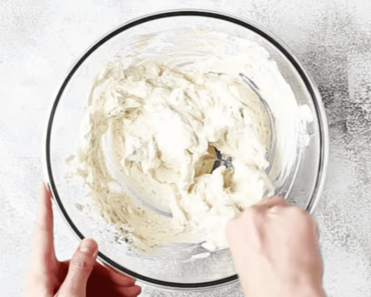 A glass mixing bowl with a creamy cheese mixture being stirred by hand.